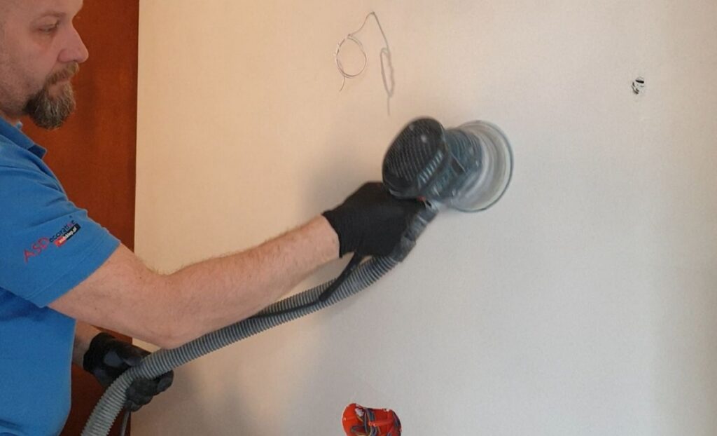 Ręczna szlifierka mimośrodowa używana do obr&oacute;bki ściany pokrytej mikrocementem. Szlifowanie mikrocementu na ścianie pomaga wygładzić powierzchnię i przygotować ją do zabezpieczenia lakierem.