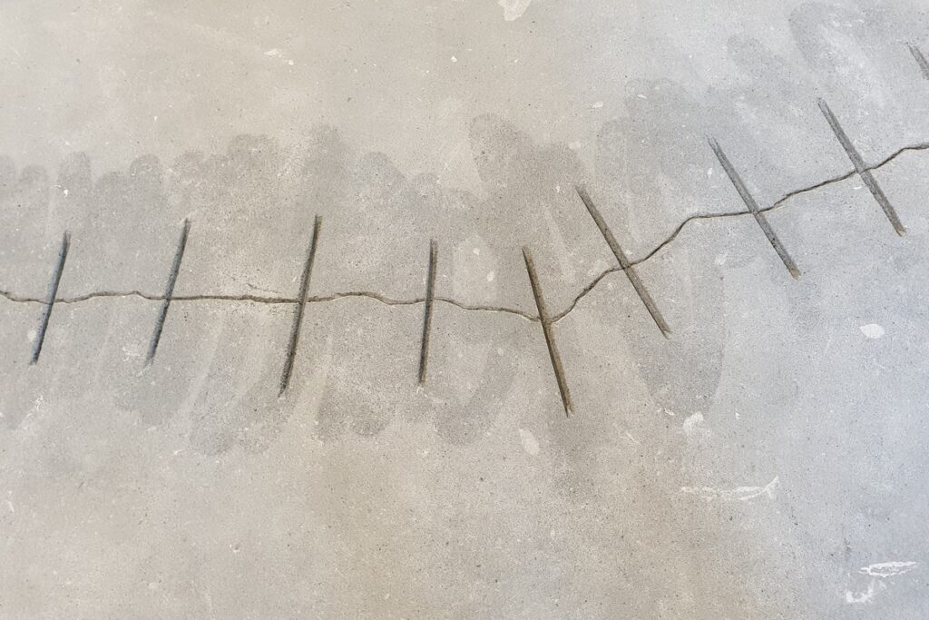 Powierzchnia podłogi z mikrocementu z widocznymi naprawionymi pęknięciami zszytymi metalowymi klamrami. To przykład przygotowania podłoża przed renowacją mikrocementu.