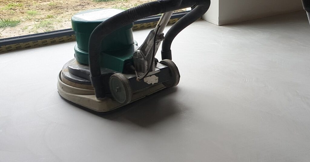 Duża szlifierka do podł&oacute;g pracująca na powierzchni z mikrocementu w pomieszczeniu. Szlifowanie mikrocementu na podłodze pozwala wyr&oacute;wnać strukturę i uzyskać jednolitą powierzchnię.