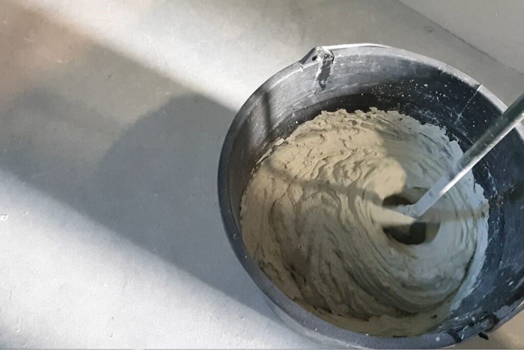 Mieszanie masy mikrocementowej w wiadrze przy użyciu mieszadła zamontowanego w wiertarce. Przygotowanie materiału jest jednym z etap&oacute;w poprzedzających zabezpieczanie powierzchni z mikrocementu.