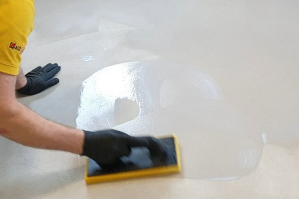 Aplikacja impregnatu na podłodze z mikrocementu przy użyciu pacy i rękawic ochronnych. Proces zabezpieczania mikrocementu zwiększa odporność powierzchni na wodę i zabrudzenia.