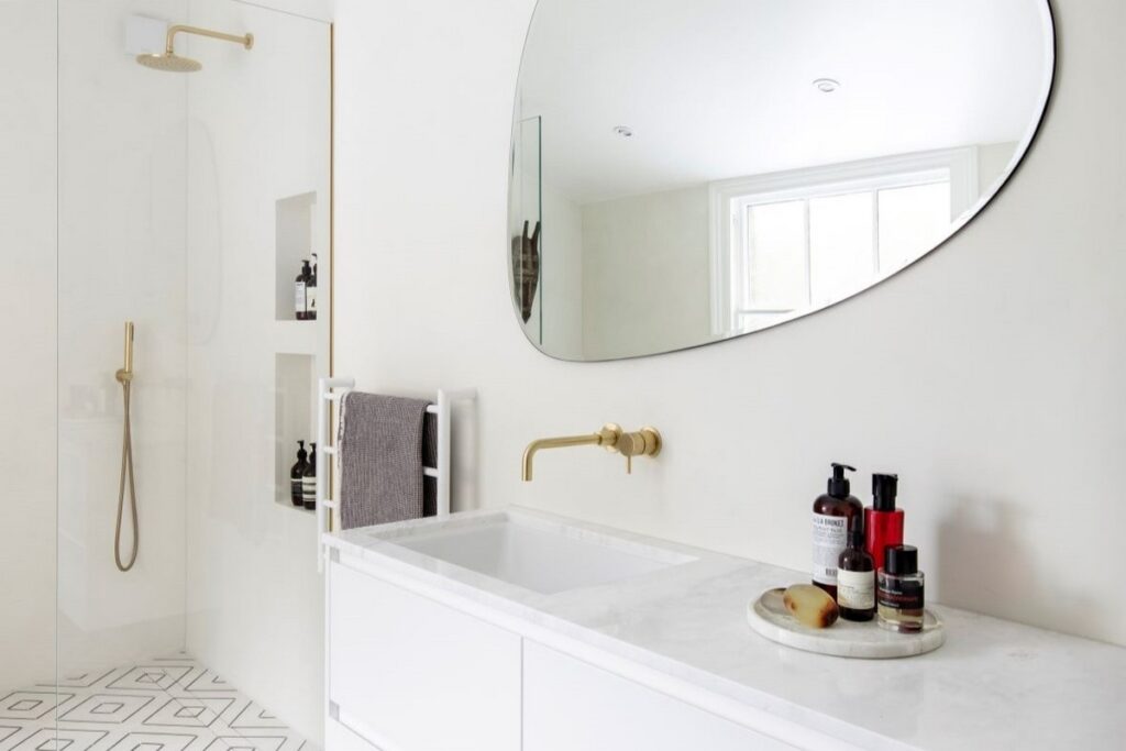 Jasna łazienka z mikrocementu z białą zabudową umywalki i dużym owalnym lustrem. Mikrocement w łazience tworzy gładkie ściany oraz strefę prysznica z armaturą w kolorze złota.