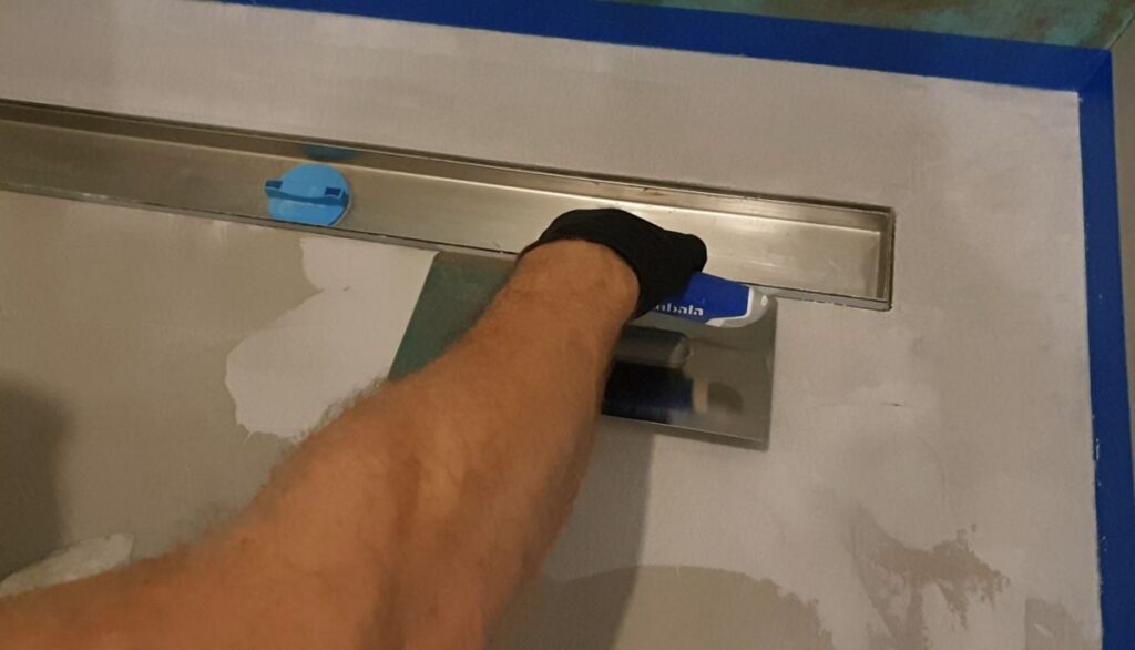Dłoń w rękawicy dociska pacą powierzchnię przy odpływie liniowym w strefie prysznica, widoczna jest taśma malarska na krawędziach. To etap, w kt&oacute;rym mikrocement w łazience wymaga precyzji przy detalach i uszczelnieniach.