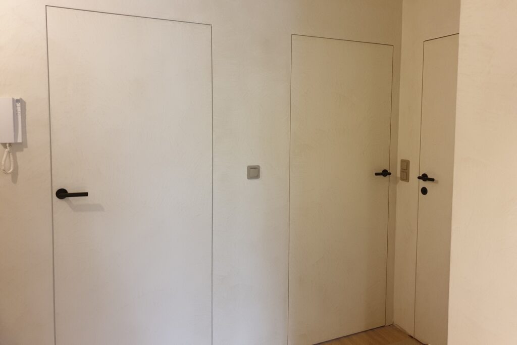 Drzwi pokryte mikrocementem zintegrowane wizualnie ze ścianą w tym samym wykończeniu. Mikrocement na meblach i drzwiach pozwala ukryć podziały w przestrzeni.