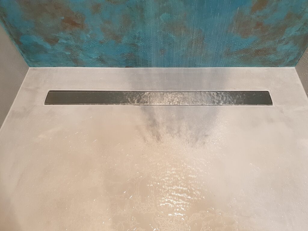 Posadzka prysznica z mikrocementu z odpływem liniowym, po powierzchni spływa woda. Mikrocement w łazience tworzy jednolitą podłogę w strefie mokrej i łączy się z kolorową ścianą w tle.