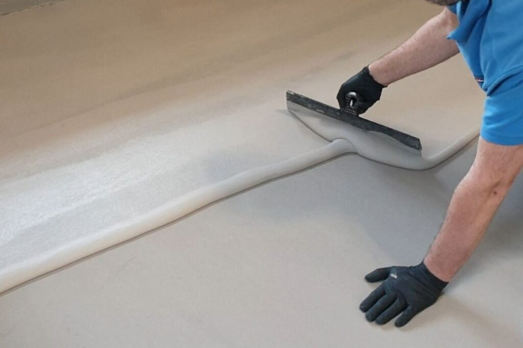 Aplikacja mikrocementu MEDIUM FAST na powierzchni przy użyciu stalowej pacy przez osobę w rękawicach. Materiał jest r&oacute;wnomiernie rozprowadzany, tworząc gładką warstwę mikrocementu FAST na podłożu.