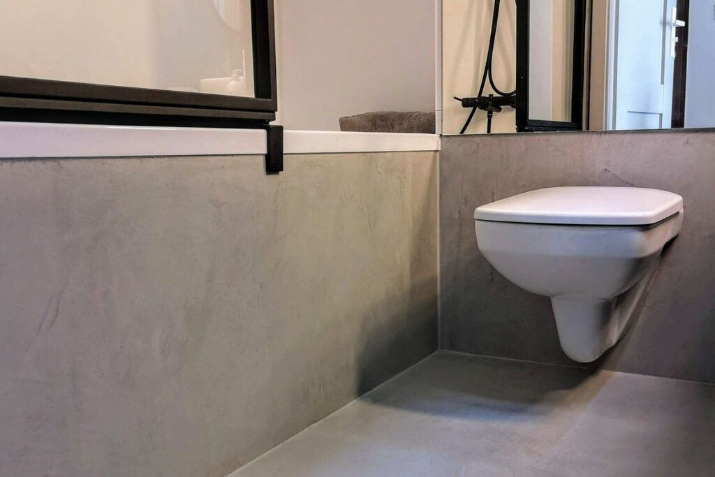Fragment łazienki z mikrocementu z szarą okładziną ścian i podłogi oraz wiszącą miską WC. Mikrocement w łazience daje jednolitą, matową powierzchnię w strefie mokrej bez widocznych spoin.