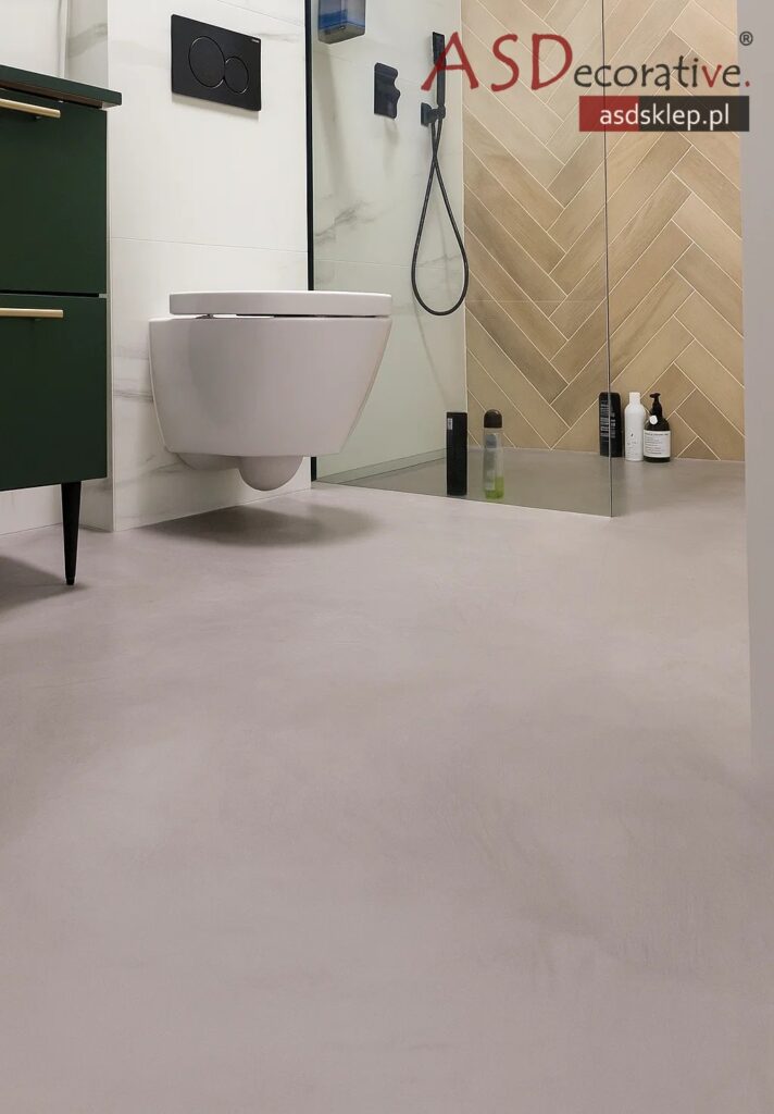 Nowoczesna łazienka z mikrocementu z szarą posadzką i wiszącą miską WC przy szklanej kabinie. W tle widać jodełkowe płytki i znak ASDecorative, a mikrocement w łazience ogranicza ilość fug.